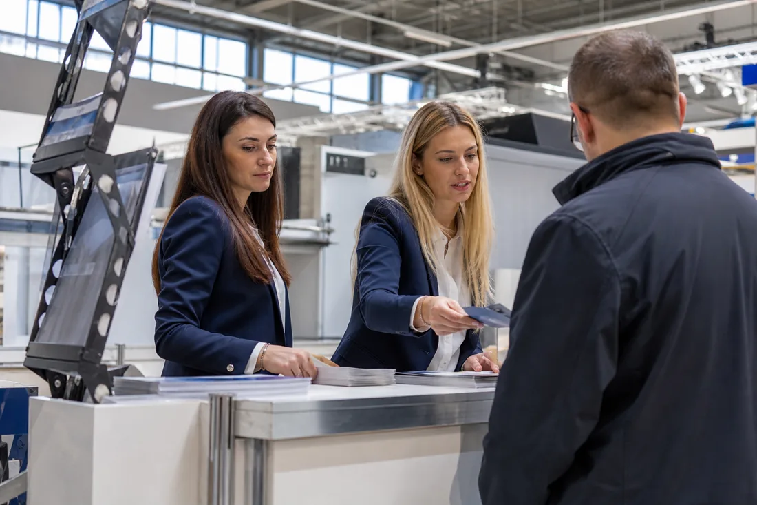 Dwie hostessy w granatowych żakietach obsługują gościa na stoisku w hali targowej